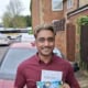 Man in a maroon shirt smiles while holding a driving certificate and magazines in front of a parked car beside a brick houseyard fence.
