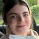 Smiling young woman in a car holds up a Practical Driving Test Pass Certificate.