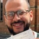 Smiling man with glasses holding a Practical Driving Test Pass Certificate outdoors by a brick building, showing his victory smile.